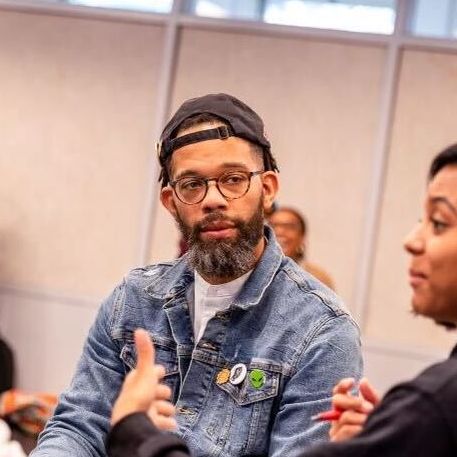 Civic Actors Studio-Cohort 5-AngieStarStudios-60_Original Photo shows Nicholas Jones participating in University of Chicago's Civic Actors Studio. He is wearing a jean jacket and a black snapback and listening to a person next to him talk.